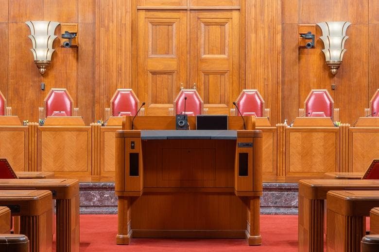 An empty hearing room with a raised wooden bench, microphones and red chairs.